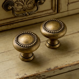 May include: Two antique brass cabinet knobs with a beaded edge and a round, domed top. The knobs are set against a distressed, light green surface, suggesting a vintage or farmhouse style. The knobs are likely used for furniture restoration or as decorative hardware.