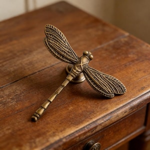 May include: A bronze-colored dragonfly drawer pull. The dragonfly has detailed wings and a textured body. The pull is mounted on a small, round base. The hardware is set on a wooden surface.