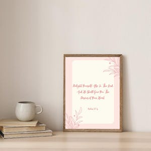 Può includere: Una stampa incorniciata con una combinazione di colori rosa chiaro e crema. Il testo recita: "Delight Yourself Also In The Lord, And He Shall Give You The Desires of Your Heart, Salmi 37:4." Una pila di libri e una tazza sono in primo piano.