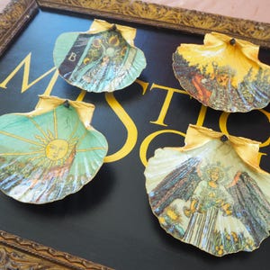 Peut inclure: Quatre coquilles Saint-Jacques décoratives avec des images de cartes de tarot, disposées sur une surface noire avec des lettres dorées. Les coquilles présentent des illustrations d'un soleil, d'un ange et d'autres figures, avec des bords peints en or.