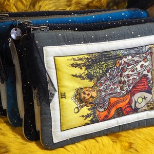 Peut inclure: Plusieurs pochettes zippées de couleurs variées, dont du bleu et du gris, ornées de paillettes argentées. La pochette de devant présente une illustration de la carte de tarot "L'Impératrice" sur fond jaune et bordure blanche. Chaque pochette a un pompon décoratif.