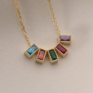 Puede incluir: Un collar dorado con una serie de gemas rectangulares de colores. Las gemas est&aacute;n dispuestas en semic&iacute;rculo, con colores como azul, rojo, verde y morado. El collar se muestra sobre una superficie blanca.