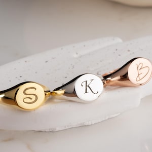 May include: Three signet rings with engraved initials. One is gold with an "S", another is silver with a "K", and the third is rose gold with a "B". The rings are displayed on a white, textured surface.