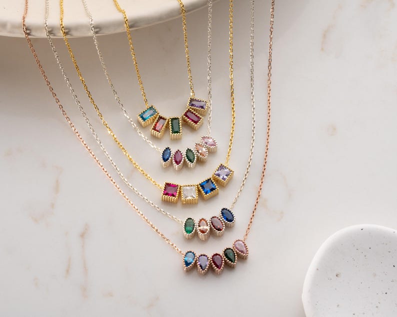 Puede incluir: Una colecci&oacute;n de delicados collares con colgantes de piedras preciosas de colores. Los colgantes presentan varias formas y colores, incluyendo azul, rosa, verde y morado. Los collares se exhiben sobre una superficie de m&aacute;rmol blanco.