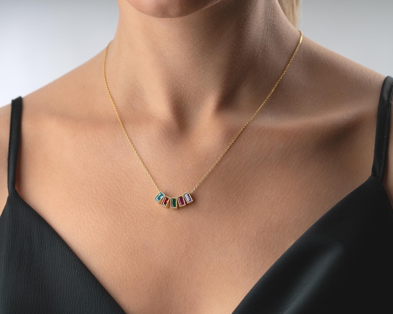 Puede incluir: Un collar dorado con una cadena fina. El collar presenta cinco piedras preciosas rectangulares de colores azul, rojo, verde y morado. Las piedras est&aacute;n dispuestas en una fila curva, creando un aspecto elegante. Una joya.