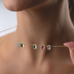 Puede incluir: Un delicado collar de oro con cinco piedras preciosas rectangulares en azul, rojo, verde, morado y transparente. Las piedras est&aacute;n engastadas en un marco dorado con cuentas y ensartadas en una fina cadena de oro. El collar est&aacute; siendo sostenido.