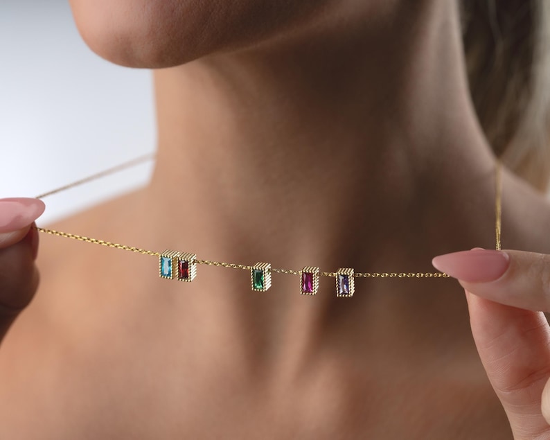 Puede incluir: Un delicado collar de oro con cinco piedras preciosas rectangulares en azul, rojo, verde, morado y transparente. Las piedras est&aacute;n engastadas en un marco dorado con cuentas y ensartadas en una fina cadena de oro. El collar est&aacute; siendo sostenido.