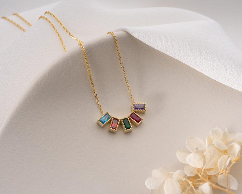 Puede incluir: Un collar dorado con una serie de gemas rectangulares de colores. Las gemas est&aacute;n dispuestas en semic&iacute;rculo, con colores como azul, rojo, verde y morado. El collar se muestra sobre una superficie blanca.