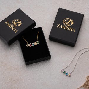 Puede incluir: Un collar dorado con cinco piedras preciosas en forma de l&aacute;grima de varios colores, exhibido en una caja negra. Tambi&eacute;n se ven dos cajas negras adicionales con texto y logotipo dorados. Tambi&eacute;n se muestra un collar plateado con piedras preciosas similares.