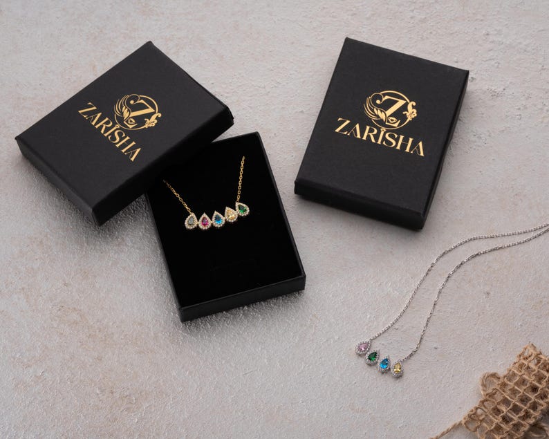 Puede incluir: Un collar dorado con cinco piedras preciosas en forma de l&aacute;grima de varios colores, exhibido en una caja negra. Tambi&eacute;n se ven dos cajas negras adicionales con texto y logotipo dorados. Tambi&eacute;n se muestra un collar plateado con piedras preciosas similares.