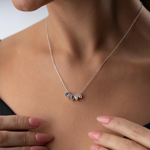 Puede incluir: Un collar de plata con una cadena fina. El colgante presenta cinco piedras preciosas de colores en forma de l&aacute;grima, azules, verdes, rosas y blancas. El collar lo lleva una persona con un top negro.