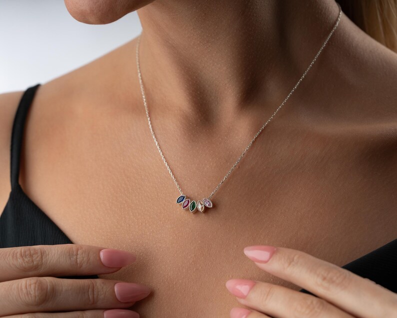 Puede incluir: Un collar de plata con una cadena fina. El colgante presenta cinco piedras preciosas de colores en forma de l&aacute;grima, azules, verdes, rosas y blancas. El collar lo lleva una persona con un top negro.