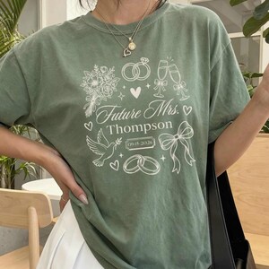 Puede incluir: Camiseta verde musgo con un diseño gráfico blanco con elementos temáticos de boda. El diseño incluye anillos, copas de champán, un ramo y el texto "Future Mrs. Thompson" con una fecha. La camiseta se combina con una falda plisada blanca.