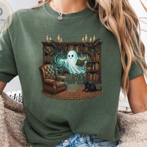 Puede incluir: Una camiseta verde musgo con un estampado gráfico de un fantasma leyendo un libro en una biblioteca con un sillón marrón, un gato negro y murciélagos. La escena incluye una estantería llena de libros, velas encendidas y una alfombra circular.