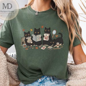 Puede incluir: Una camiseta de color verde oliva oscuro con una ilustración gráfica de cuatro gatos negros realizando diversas actividades. Los gatos aparecen leyendo libros, sosteniendo un portapapeles con la etiqueta IEP e interactuando con un espejo y una taza.