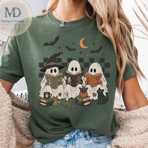 Puede incluir: Una camiseta de color verde oliva oscuro con un gráfico de tres fantasmas leyendo libros. El diseño incluye una luna creciente, murciélagos, un gato negro, pilas de libros, una vela y una calabaza. Los fantasmas llevan sombreros y libros abiertos.