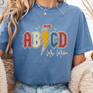 Puede incluir: Una camiseta azul con un diseño gráfico que muestra las letras AB y CD en una fuente roja y blanca, separadas por un rayo amarillo con forma de lápiz. Debajo del gráfico, el texto Mrs. Miller está impreso en una fuente amarilla cursiva.