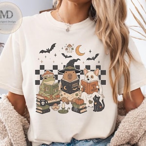 Puede incluir: Una camiseta de color marfil con un estampado gráfico de una rana, un capibara con sombrero de bruja y un perro disfrazado de fantasma, todos leyendo libros. El diseño incluye murciélagos, una luna creciente, setas, una calabaza y un gato negro.