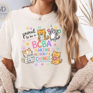 Puede incluir: Una camiseta color hueso con un gráfico colorido de cuatro gatos de dibujos animados y el texto "proud to be a BCBA MAKING MEANINGFUL CHANGE". El diseño incluye pequeñas flores, corazones y estrellas, con dos gatos sosteniendo un horario visual.
