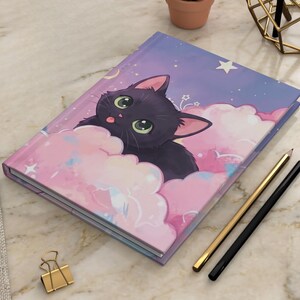 Puede incluir: Un cuaderno de tapa dura con una ilustración de un gato negro de grandes ojos verdes y lengua pequeña asomando, descansando entre nubes rosas y azules sobre un cielo estrellado violeta. Incluye dos lápices y un clip de metal dorado.
