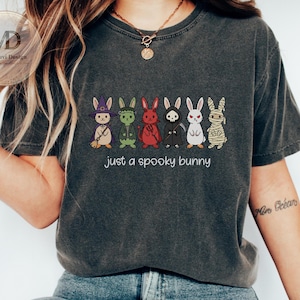 May include: A charcoal gray t-shirt featuring a graphic of six cartoon bunnies dressed in Halloween costumes, including a witch, Frankenstein, devil, grim reaper, vampire, and mummy, with the text "just a spooky bunny" printed below.