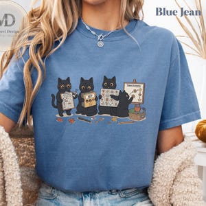 Puede incluir: Una camiseta azul con un gráfico de cuatro gatos negros realizando actividades escolares. Los gatos sostienen un portapapeles, una carpeta IEP, un horario visual y un tablero de economía de fichas con estrellas, lápices y hojas.