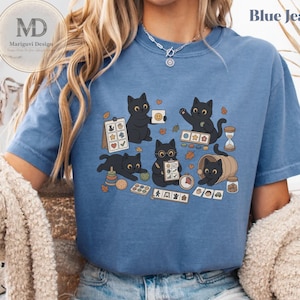 Puede incluir: Una camiseta azul con un estampado gráfico de cinco gatos negros realizando diversas actividades con juguetes, incluyendo un portapapeles, un reloj de arena y un juguete apilable. El diseño incluye pequeñas hojas de otoño y el texto Blue Jean.