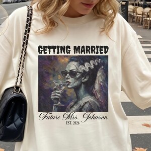 Puede incluir: Una camiseta de color marfil con un estampado gráfico de una novia con pelo oscuro y gafas de sol sosteniendo una copa de martini. El texto GETTING MARRIED aparece sobre la imagen y Future Mrs. Johnson EST. 2026 debajo en letras negras.