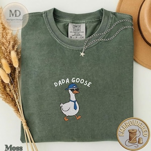 Könnte beinhalten: Ein moosgrünes T-Shirt mit Rundhalsausschnitt und einer Stickerei einer weißen Gans, die eine blaue Baseballkappe, eine Sonnenbrille und eine blaue Krawatte trägt. Der Text DADA GOOSE ist in Weiß darüber gestickt. Mit Comfort Colors Etikett.