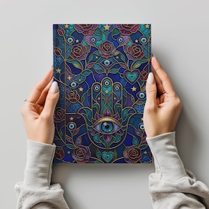 Puede incluir: Un diario decorativo con un diseño de vidriera que presenta una mano de Hamsa, rosas y ojos. La cubierta es predominantemente azul con detalles dorados, y el diseño incluye elementos morados, turquesas y dorados. El diario es sostenido por una persona.
