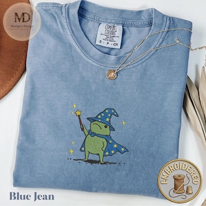 Puede incluir: Camiseta azul vaquero con un diseño bordado de un mago rana verde sosteniendo una varita. La rana lleva una capa y un sombrero azules con estrellas amarillas. La camiseta tiene una etiqueta Comfort Colors y un pequeño collar dorado.