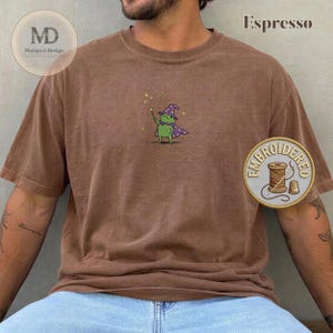 以下が含まれることがあります： エスプレッソブラウンの半袖Tシャツ。紫色の魔法使いの帽子とマントを身につけ、黄色い星がついた杖を持つ緑色のカエルの小さな刺繍が施されています。クルーネックでゆったりとしたフィット感のTシャツです。