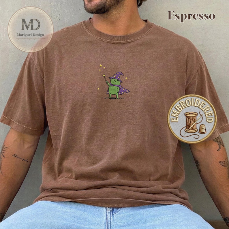 以下が含まれることがあります： エスプレッソブラウンの半袖Tシャツ。紫色の魔法使いの帽子とマントを身につけ、黄色い星がついた杖を持つ緑色のカエルの小さな刺繍が施されています。クルーネックでゆったりとしたフィット感のTシャツです。