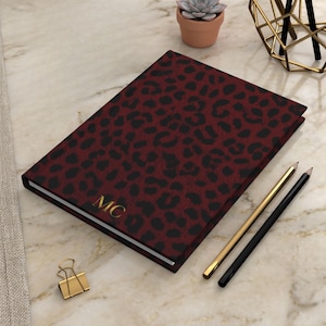 May include: A closed journal with a burgundy leopard print cover and gold initials "MC". Two pencils, one gold and one black, are next to the journal. A gold paper clip and a small potted succulent are also visible.