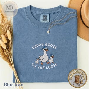 May include: A blue short-sleeved t-shirt featuring an embroidered graphic of a large goose and two smaller geese, all wearing blue baseball caps and sunglasses. The text DADDY GOOSE ON THE LOOSE is embroidered in white arched lettering around the geese.