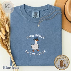 Könnte beinhalten: Ein blaues Kurzarm-T-Shirt mit Rundhalsausschnitt und einer Stickerei einer weißen Gans, die eine blaue Baseballkappe und eine schwarze Sonnenbrille trägt, mit dem Text DADA GOOSE ON THE LOOSE in weißen Buchstaben.