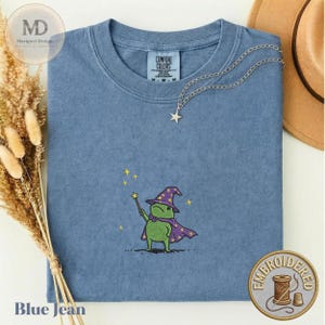 以下が含まれることがあります： 紫色の魔法使いの帽子とマントを身につけ、星の杖を持った緑色のカエルの刺繍が施された青いクルーネックTシャツ。シャツの首元にはComfort Colorsのラベルがあり、銀色の星のペンダントネックレスが添えられています。