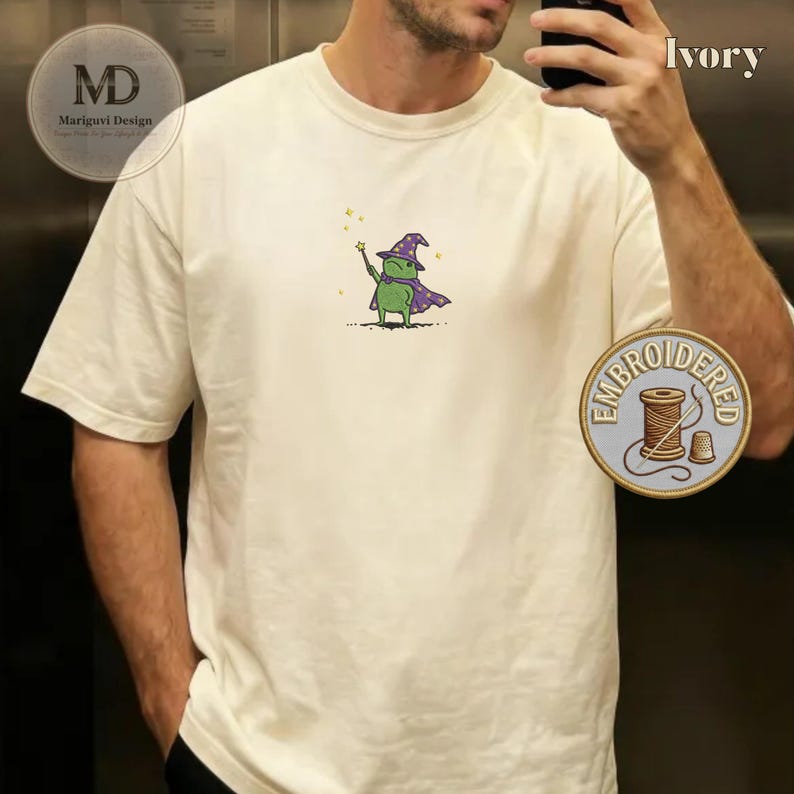 以下が含まれることがあります： アイボリー色の半袖Tシャツ。胸元には、紫色の魔法使いの帽子とマントを身につけ、星のついた杖を持った緑色のカエルの小さな刺繍が施されています。デザインには、カエルの周りに小さな黄色い星が散りばめられています。