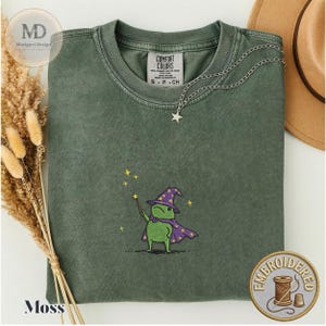 以下が含まれることがあります： モスグリーンのクルーネックTシャツ。紫色の魔法使いの帽子とマントを身につけ、黄色い星がついた杖を持つ緑色のカエルの刺繍が施されています。TシャツにはComfort Colorsのラベルが付いています。