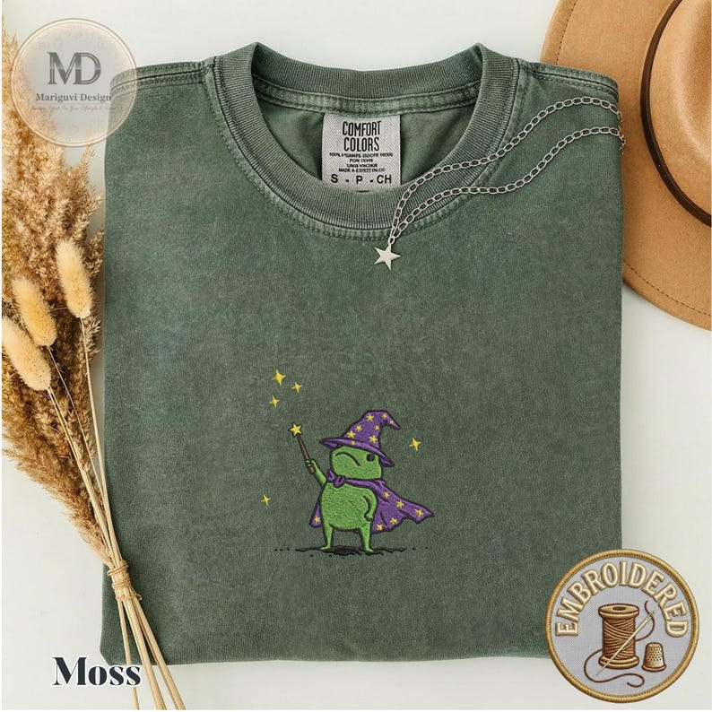 以下が含まれることがあります： モスグリーンのクルーネックTシャツ。紫色の魔法使いの帽子とマントを身につけ、黄色い星がついた杖を持つ緑色のカエルの刺繍が施されています。TシャツにはComfort Colorsのラベルが付いています。