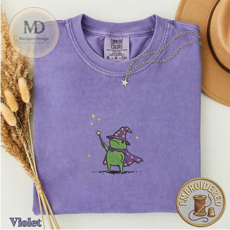 以下が含まれることがあります： 紫色のクルーネックTシャツ。紫色の魔法使いの帽子とマントを身につけ、星の杖を持った緑色のカエルの刺繍が施されています。首元にはComfort Colorsのラベルがあり、銀色の星型ペンダントネックレスが添えられています。