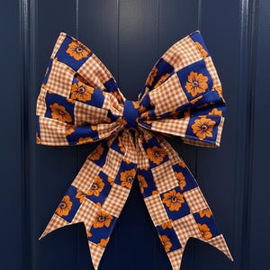 Op de afbeelding: Een grote decoratieve strik met een blauw en oranje bloemen- en ruitpatroon. De strik is gemaakt van stof en heeft een centrale knoop en lange, puntige uiteinden. De strik hangt aan een donkerblauwe deur.