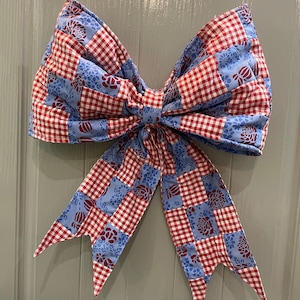 May include: A large, decorative bow with a patchwork design. The bow features a combination of red and white gingham check fabric and blue fabric with a floral pattern. The bow is likely used for decoration, possibly on a wreath or gift.