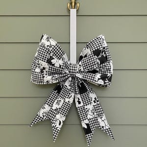 May include: A large decorative bow with a black and white floral and gingham pattern. The bow is hanging from a decorative gold hook with a white ribbon. The bow is made of fabric and is likely used for home decor.
