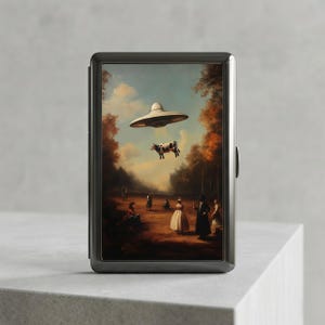 May include: A black metal cigarette case featuring a surreal painting of a cow being abducted by a UFO over a field with people. The artwork has a vintage, classical style with a blue sky and trees.