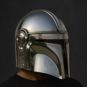 Pu&ograve; includere: Un lucido elmo Mandaloriano argentato, con una caratteristica visiera a forma di T e dettagli laterali intricati. L'elmo &egrave; indossato da una persona con una maglietta nera, su uno sfondo scuro. Questa &egrave; una replica della serie di Star Wars.