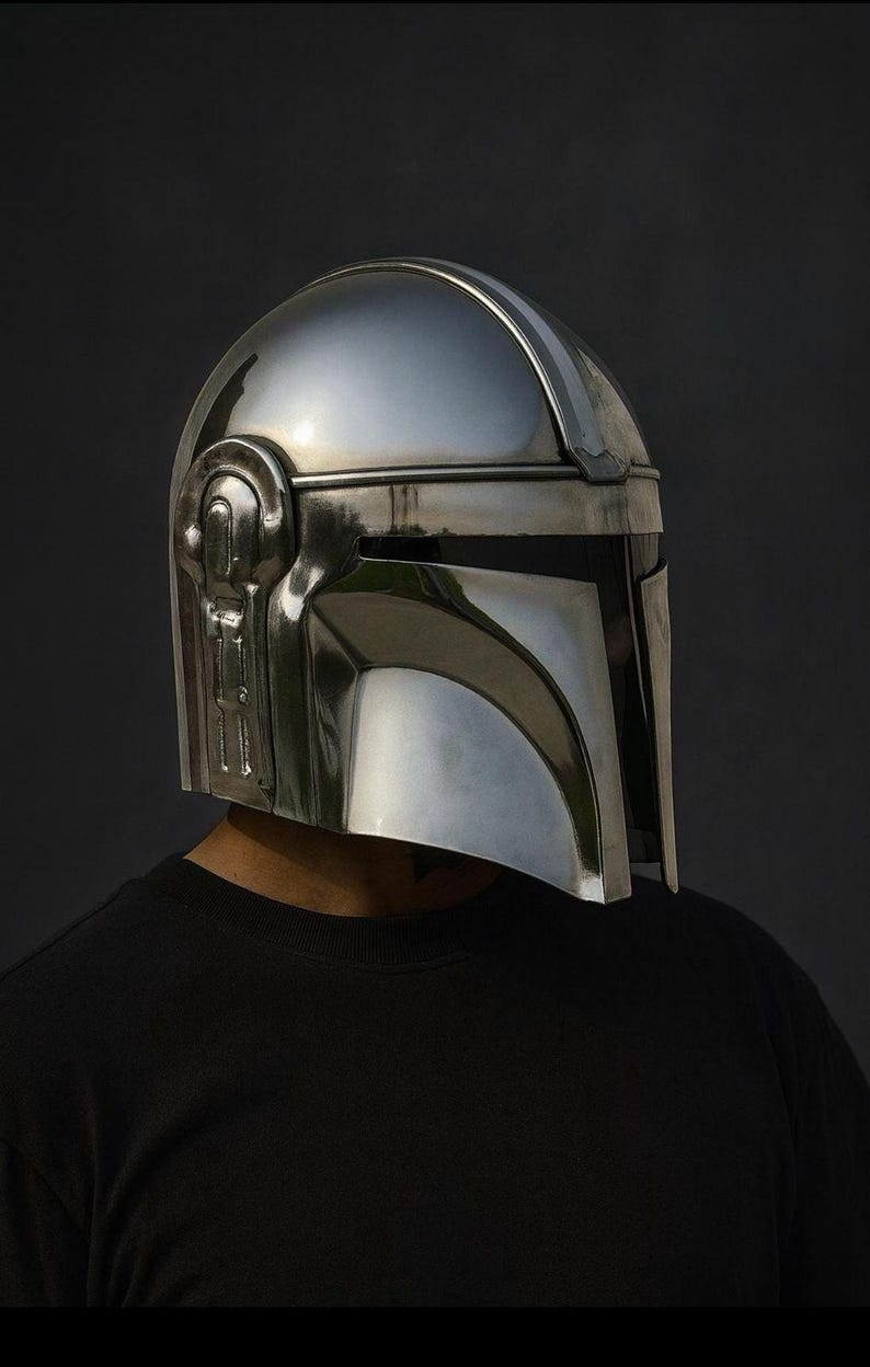 Pu&ograve; includere: Un lucido elmo Mandaloriano argentato, con una caratteristica visiera a forma di T e dettagli laterali intricati. L'elmo &egrave; indossato da una persona con una maglietta nera, su uno sfondo scuro. Questa &egrave; una replica della serie di Star Wars.