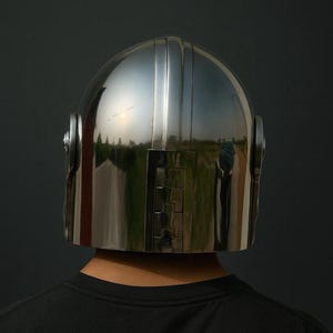 Pu&ograve; includere: Un casco argentato lucido con una parte superiore arrotondata e scanalature verticali. Il casco presenta una sezione rettangolare nella parte anteriore e dettagli circolari sui lati. La superficie riflettente mostra uno sfondo scuro.