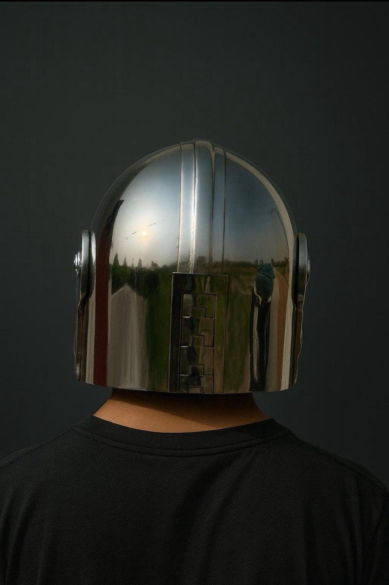 Pu&ograve; includere: Un casco argentato lucido con una parte superiore arrotondata e scanalature verticali. Il casco presenta una sezione rettangolare nella parte anteriore e dettagli circolari sui lati. La superficie riflettente mostra uno sfondo scuro.