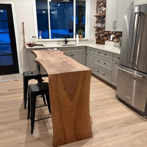 May include: A bespoke kitchen island with a live edge wooden worktop. The island is crafted from light brown wood and has a natural, organic form. Two black metal stools are placed in front of the island. The kitchen features grey cabinets and stainless steel appliances.